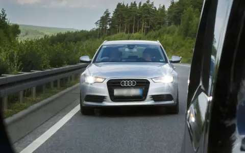 Silberner Audi auf der Autobahn, blendet mit Lichthupe. Vorbei an grünen Wäldern und einer Leitplanke.
