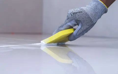 Mano enguantada aplica masilla blanca para rejuntar baldosas con una espátula amarilla.