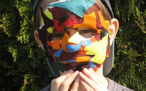 Dziecko w kolorowej masce z filcu, symbolizującej maskowanie autyzm. Dłonie przy ustach.