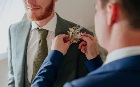 Un homme ajuste une boutonnière sur le costume d'un autre. Divers types de boutonnières existent, celle-ci est composée de fleurs séchées et de feuillages dorés.