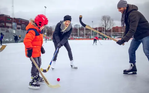 Familj njuter av ishockey i Göteborg. En pappa, mamma och son med hockeyklubbor och en röd puck på isen.