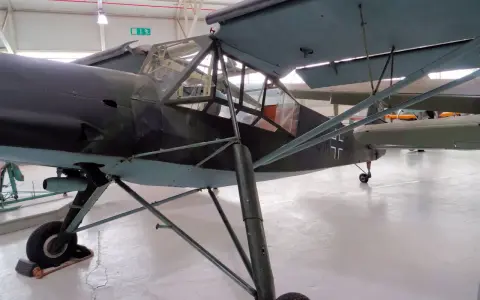 Ein altes Flugzeug, ein Storch, steht in einer Halle. Das Flugzeug hat eine Tarnlackierung und ein Kreuz auf dem Heck.