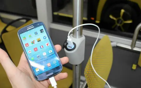 Dłoń trzyma smartfon podłączony do ładowarki USB zamontowanej na słupku w autokarze.