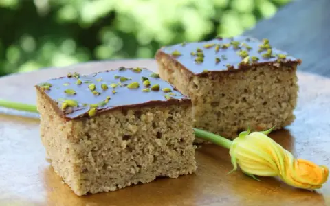 Zwei Stücke saftiger Zucchini Kuchen mit Schokoladenglasur und gehackten Pistazien, daneben eine Zucchiniblüte.