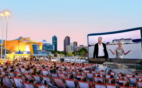 Ein lauer Sommerabend im Freiluftkino Charlottenburg. Menschen sitzen in Liegestühlen und schauen auf eine Leinwand, auf der ein Film mit einem älteren Mann und einer jungen Frau läuft. Im Hintergrund die Berliner Skyline.