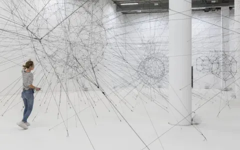 Une femme admire une installation de Tomas Saraceno, un réseau complexe de fils noirs tendus dans un espace blanc.