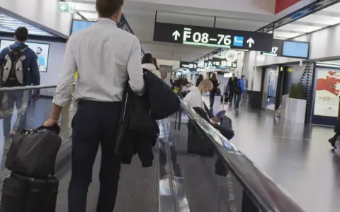 Resenärer på rullband på flygplatsen, skyltar visar vägen till gate F08-76. En man med resväskor på väg mot centrum.