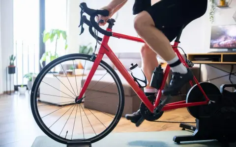 Intensywny trening na trenażerze kolarskim w domu. Czerwony rower, sportowiec w spodenkach i koszulce, butelki z wodą.