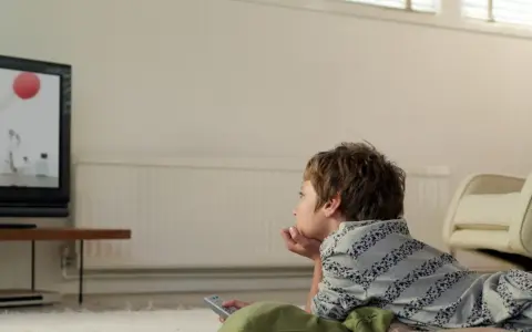 Chłopiec leży na dywanie przed telewizorem, oglądając bajkę. Trzyma pilota, gotowy do zmiany kanału.