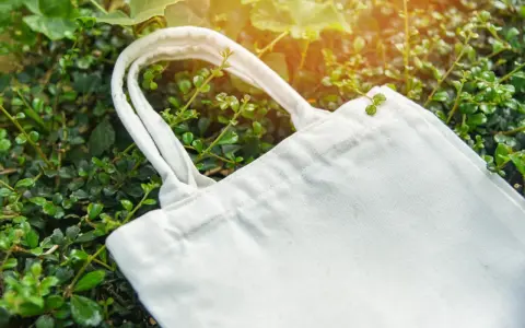 Eine weiße Stofftasche liegt zwischen grünen Blättern. Perfekt, um die Einkaufsstaschen zu verstauen und die Umwelt zu schonen.