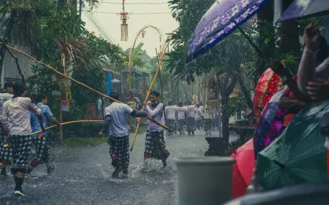 Ulewny deszcz na Bali w lutym. Mężczyźni w tradycyjnych strojach niosą drewniane tyczki, a widzowie chronią się pod kolorowymi parasolami.