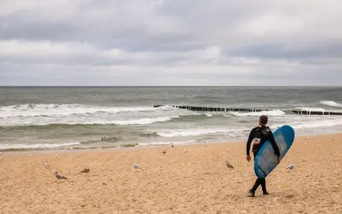 Surfer idzie plażą w Juracie, szukając fal. To jedna z wielu atrakcji tego miejsca.