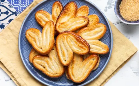 Un piatto blu pieno di dolci con pasta sfoglia dorati e croccanti, cosparsi di zucchero.