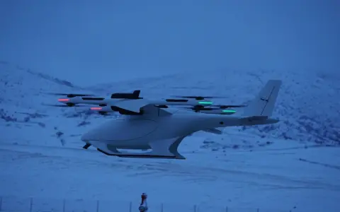 A white vtol drone with red and green lights flies over a snowy landscape at dusk.