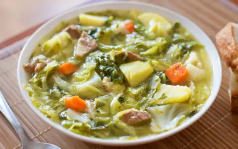 Un bol de soupe aux choux réconfortante, avec des morceaux de chou, pommes de terre, carottes et viande. Du pain frais à côté.