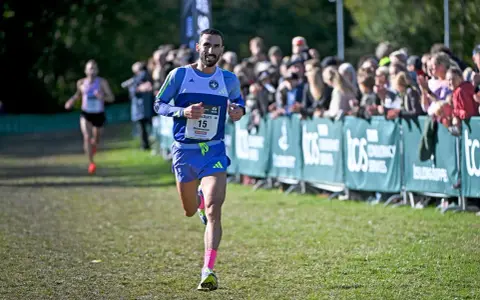 Löpare med nummer 15, klädd i blått och med rosa strumpor, springer mot publiken. En annan löpare syns i bakgrunden. Kanske är det en del av sevilla marathon.