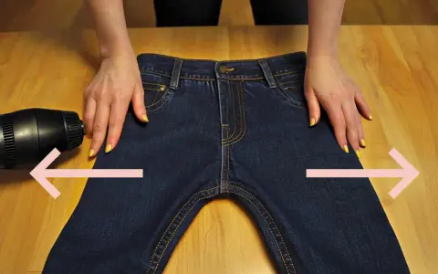 Manos sujetan pantalones vaqueros oscuros sobre mesa de madera. Secador de pelo apunta a la tela. Aprende como estirar pantalones vaqueros.
