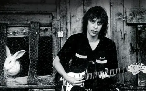 Ein Mann mit Gitarre, im Hintergrund ein Hase im Käfig. Ein Bild, das an den **titel von Rio Reiser** erinnert.
