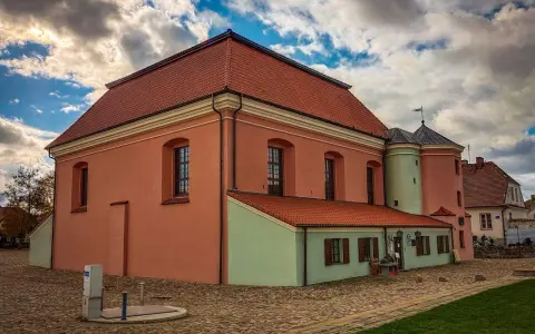 Tykocin co zobaczyć: zabytkowa Wielka Synagoga z różową fasadą i zielonym aneksem, na brukowanym placu pod burzliwym niebem.
