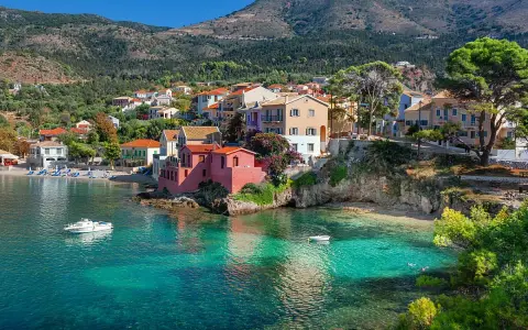 Case colorate affacciate su acque cristalline, un sogno di spiagge Grecia. Barche a vela e colline verdi completano il quadro.