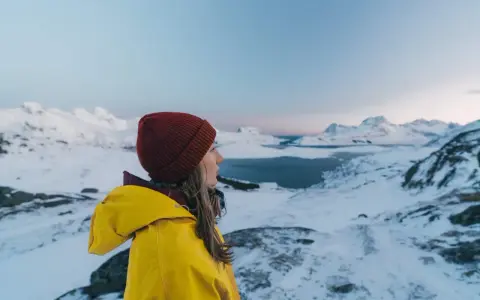 Kvinna i gul jacka och röd mössa njuter av utsikten över snöklädda berg och hav. Perfekt för sportlov i Norge.