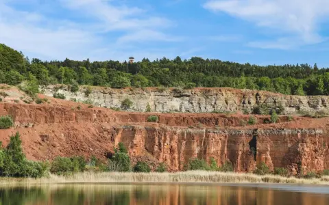 En sjö speglar Sveriges Grand Canyon, en imponerande klippformation med lager av röd och grå sten, krönt av en grönskande skog.