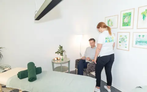 En kvinna i vit tröja med texten "HÄLSO RESURS" pratar med en man som sitter i en fåtölj. Rummet har en massagebänk och konst på väggarna, kanske för att förbereda för en vagusnerven massage.