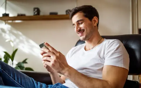 Uśmiechnięty mężczyzna w białej koszulce siedzi w fotelu i patrzy na telefon.