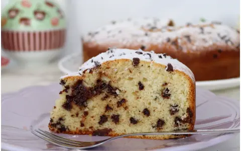Una fetta di torta soffice con tante gocce di cioccolato, spolverata di zucchero a velo, su un piatto lilla con una forchetta.
