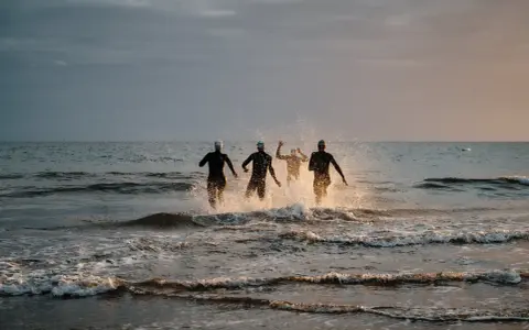 Vier Schwimmer stürmen ins Meer. Die durchschnittliche Zeit für 1km schwimmen wird hier wohl im Wettkampf ermittelt.