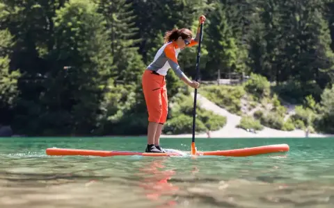 Kobieta w pomarańczowych spodenkach i sportowej bluzce uprawia pływanie na supie po spokojnym jeziorze. W tle zielone drzewa i jasna plaża.