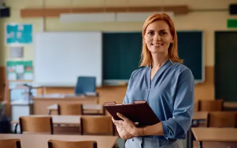 Uśmiechnięta nauczycielka z rudymi włosami trzyma teczkę. W tle klasa z ławkami i tablicą. Posiada legitymację nauczyciela.