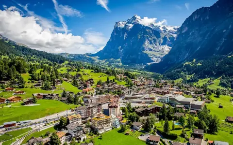 A scenic view of Grindelwald, Switzerland, nestled in a valley with lush green hills and a majestic snow-capped mountain. A train travels through the village.
