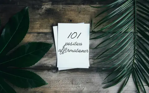 En hög med papper med texten "101 positiva affirmationer" på en träbakgrund, omgiven av gröna blad. En vacker påminnelse om affirmation svenska.