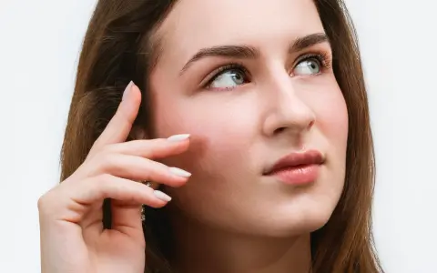 Une jeune femme aux yeux bleus regarde au loin, sa main effleurant sa joue. Ses cils, sublimés par un **tatouage cils**, accentuent son regard captivant.