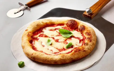 Frisch gebackene Pizza Margherita mit geschmolzenem Mozzarella und Basilikum. Daneben liegen ein Pizzaschneider und eine Pizzaschaufel, bereit, den perfekten pizzateig ausrollen.