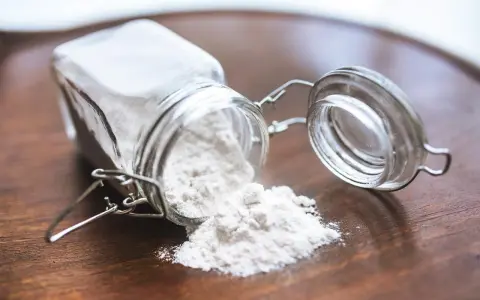 Un barattolo di vetro rovesciato su un tavolo di legno, con polvere bianca che fuoriesce, forse ingredienti per colla vinilica fatta in casa.