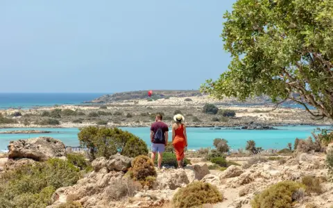 Ett par njuter av utsikten över Elafonissi, Kreta, med dess turkosa vatten och steniga landskap. En röd fallskärm syns i fjärran.