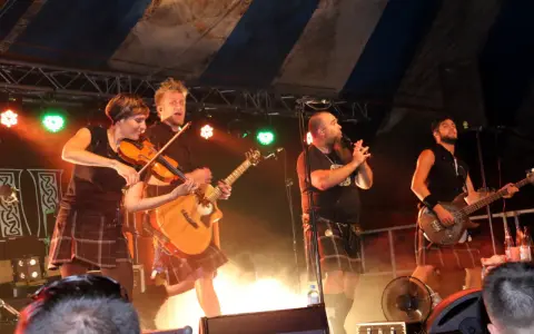 Un groupe celtique français sur scène, avec violon, guitare et basse. Les musiciens portent des kilts, créant une ambiance festive.