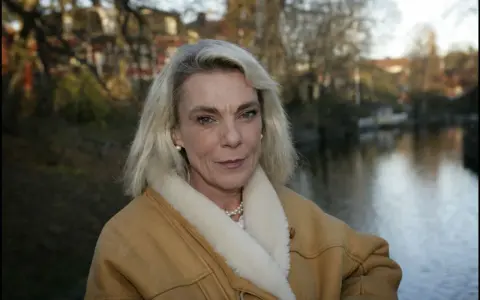 Pia Degermark idag, med sitt blonda hår och en varm fårskinnsjacka, står vid en vattensamling med träd och hus i bakgrunden.