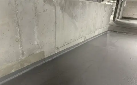 Szara, gładka posadzka w garażu, zabezpieczająca co na podłogę w garażu przed wilgocią. Betonowa ściana w tle.