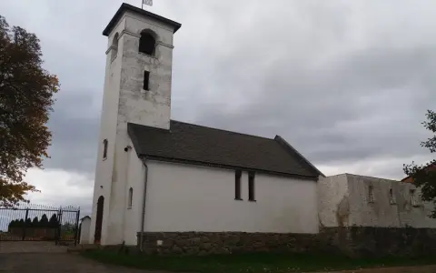 Biały, zabytkowy dwór z wieżą i dachem krytym dachówką. W oddali widać bramę i drzewa.