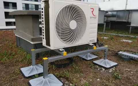 Jednostka zewnętrzna klimatyzatora Rotenso na dachu, z widocznymi kanałami wentylacyjnymi i budynkiem w tle.