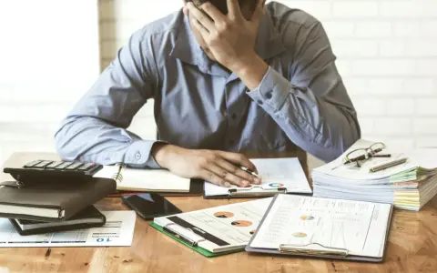 Zestaw dokumentów, kalkulator i telefon leżą na biurku. Mężczyzna z ręką na czole, przytłoczony problemami finansowymi, przegląda pismo windykacyjne.