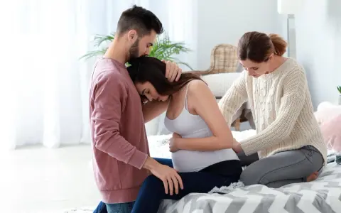 Mężczyzna obejmuje ciężarną kobietę, która opiera głowę na jego ramieniu. Obok siedzi doula, która delikatnie dotyka jej brzucha, oferując wsparcie.