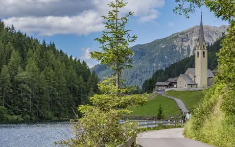 Passeggiata sul lago di Valdurna, con boschi rigogliosi, una chiesa alpina e montagne sullo sfondo.