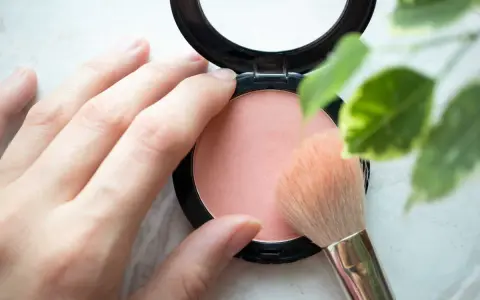 Une main applique un pinceau sur un blush poudre, révélant une douce teinte couleur blush.