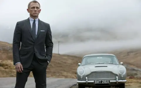 Uomo con impeccabile 007 outfit, completo gessato e cravatta, posa accanto a una Aston Martin d'epoca in un paesaggio nebbioso.