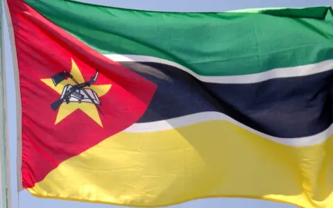 Flaga Mozambiku: zielono-czarno-białe pasy, czerwony trójkąt z żółtą gwiazdą, karabin AK-47 i książka.