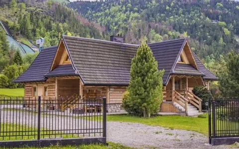 Drewniana bacówka pod skocznią, otoczona zielenią, z widokiem na górskie zbocza.